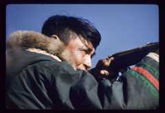 A colour photograph of a man seen in profile aiming a gun. He’s wearing a traditional fur-trimmed parka with alternating green and red stripes on the sleeves.