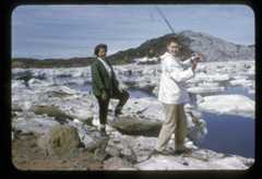 A colour photograph of two women fishing on the banks of a water body. They are standing on rocks and there are ice floes in the water. 