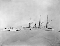 Black-and-white photo of a ship surrounded by snow and ice, with people next to it building a snow shelter.