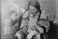 Black-and-white photo of a woman sewing skin boots, while a child plays with her braids.