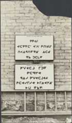 Photograph of a close-up view of a light grey brick wall. There are two rectangular white signs attached to the centre of the wall. The upper sign has four lines of syllabics, and the lower one has five lines of syllabics. Both are in black ink. There is a ladder laying horizontally across the base of the wall.