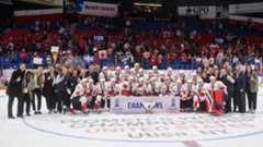 Female hockey players posing with their gold medals, championship banner and trophy on an ice rink.