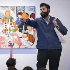 A museum educator stands in front of a colorful artwork depicting a lively dinner scene. The educator, wearing glasses, a beard, a blue jacket, and carrying a tote bag, gestures and points while engaging a group of children seated out of frame, one of whom raises their hand to ask a question.