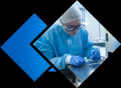 A person in a blue surgical gown, gloves, and hairnet carefully examines a metallic object on a stainless steel surface in a lab setting.
