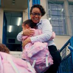 An adult wearing glasses hugs a child with a pink backpack outside a building with blue railings and windows.