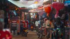 A bustling market scene with colorful fabric canopies overhead, motorbikes parked along the dirt pathway, and people walking and shopping among stalls selling clothes and household goods.