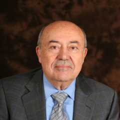Andrew Viterbi, an elderly man with a bald head, wearing a blue dress shirt, gray checkered suit jacket, and a gray tie, posing against a dark brown background.