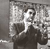 Teller with a molecule model in front of the periodic table giving a lecture.