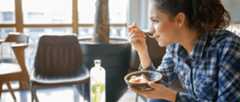 A woman sitting on a couch eating breakfast from a bowl in her hand while her coffee and bottle of water sit on the table in front of her.