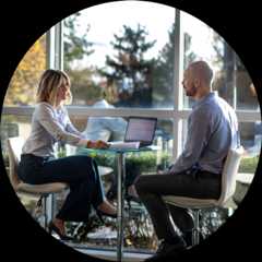 A man and woman work colleagues sitting at a table by a large window reviewing work on a laptop and printed-out papers.
