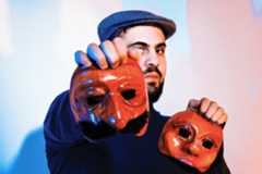 A Palestinian man wearing a flat cap and black jumper holds up two masks, one of which slightly obscures his face.