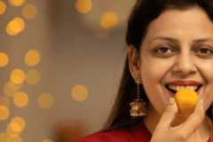 A South Asian woman wearing a red top and gold earrings holds her hand to her mouth whilst eating.