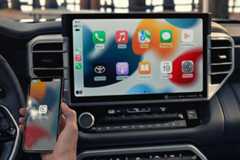 A hand holding an iPhone displaying the CarPlay icon, positioned in front of a cars dashboard screen which shows the Apple CarPlay interface with various app icons.