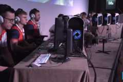 A group of young men wearing headphones and matching shirts are seated at a long table with computers, appearing to be engaged in an esports competition.