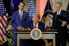 Donald Trump signing a document at a podium, flanked by two men and American flags.