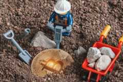 A Lego miner figure using a jackhammer next to a partially buried golden Bitcoin, with a shovel and a wheelbarrow full of rocks, all on a dirt ground.