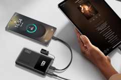 A smartphone and a tablet are being charged by a black power bank. The smartphone screen shows 80% battery, and the tablet displays a music player interface. A hand holds the tablet.