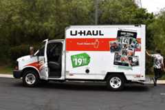 A U-Haul truck with its door open, parked on asphalt with trees in the background. A person is standing near the back of the truck.