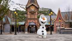 A full-body Olaf mascot stands on a cobblestone path in front of a Frozen themed village with snow-capped mountains in the background.