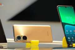 A gold smartphone is displayed horizontally on a yellow stand, with its dual cameras visible. To its right, a black smartphone stands vertically, displaying a green and blue interface with the time 10:31.