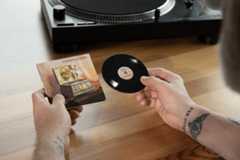 A person holding a small vinyl record and its album art, with a turntable in the background, resized to a 16:9 aspect ratio.