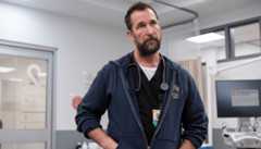 A man with a beard wearing a dark blue hoodie over scrubs and a stethoscope around his neck, standing in what appears to be a hospital room.
