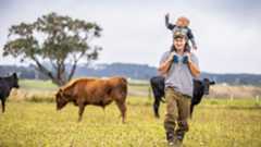 man carrying boy on shoulders in paddock with cow in background 