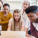 A group of young ASX investors sitting around a laptop with an older lady standing behind them explaining how investing works.