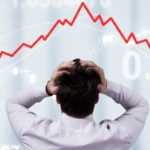 A man with his back to the camera holds his hands to his head as he looks to a jagged red line trending sharply downward.