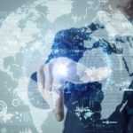 a man in a business suit points his finger amid a digitised map of the globe suspended in the air in front of him, complete with graphs, digital code and glyphs to indicate digital assets.