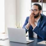 A male investor wearing a white shirt and blue suit jacket sits at his desk looking at his laptop with his hands to his chin, waiting in anticipation.