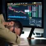A man lays his head down on his arms at his desk in front of an array of computer screens and a laptop computer.