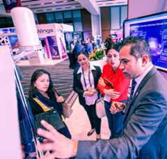 A group of four people, three women and a man, attentively observe and discuss a presentation on a large screen. The setting appears to be a busy convention center, with branded booths and a "Wings" sign visible in the background. The group is dressed in business attire.