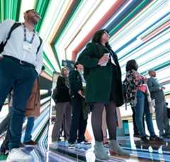 A group of people stand indoors, looking up at a vivid, multicolored LED light installation on the ceiling. The light beams create a dynamic, futuristic atmosphere. Most people wear business attire with conference badges, suggesting an event or exhibition.