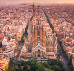 Aerial view of the Sagrada Família basilica in Barcelona at sunset, surrounded by city blocks. The iconic church features intricate spires and ongoing construction with cranes visible. The cityscape extends into the hazy distance under a pink-orange sky.