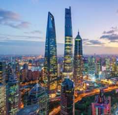 A vibrant cityscape at dusk shows Shanghai’s skyline with the Shanghai World Financial Center and Shanghai Tower rising above illuminated skyscrapers, glowing streets, and the Huangpu River under a partly cloudy sky.
