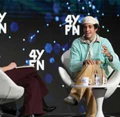 Two people sit in white chairs on a stage with a "4YFN" logo backdrop. The person on the right, wearing a white hat, mint sweater, and khaki pants, gestures while speaking. A carafe and glasses with the "4YFN" logo sit on a small table between them.