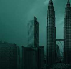 A misty cityscape featuring the tall, twin Petronas Towers with their skybridge in Kuala Lumpur, Malaysia. Modern high-rise buildings surround the towers, and the overall image has a teal color tone.