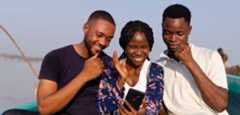Three people sit closely together outdoors in a boat, smiling and looking at a smartphone held by the woman in the center. The background shows calm water and a clear sky. They appear happy and engaged with what they see on the phone.