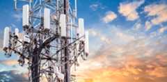 A close-up view of a telecommunications tower with multiple white antennas and cables, set against a vibrant sky with scattered clouds and a warm, colorful sunset in the background.