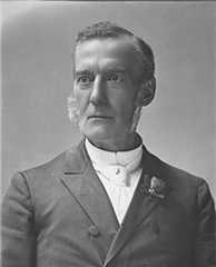 Head and shoulders portrait of a man with sideburns, wearing a suit coat, high collared shirt, and white tie. He looks off to the side, and has a small flower pinned to his lapel.  