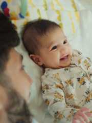 花柄のロンパースを着た黒髪の赤ちゃんが仰向けになって笑っていて、それを父親が見つめている。