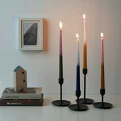 Vier kleurrijke dinerkaarsen in bruin, blauw, groen en beige met vervagende kleuren, rechtopstaand in zwarte kaarsenhouders.