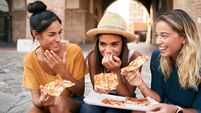 Three beautiful women sitting on the stairs of the city streets eating pizza from a street stall. The happy girls enjoy the week