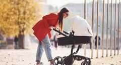 Mother in red coat have a walk with her kid in the pram in the park at autumn time