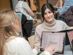 Two people sit at a table talking with laptops open in front of them.