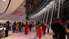 A group of people gather in the modern, glass-walled atrium with tall white columns of the Kansas City Performing Arts center. Some are dressed formally, others casually, while a few engage in conversation and others walk around.