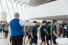 A man in a blue vest gestures toward a group of students in uniforms walking inside the large, modern Kauffman Center with its curved ceiling and expansive windows.