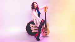 A woman with a white leather jacket sits on a bass drum that says "Danielle Nicole Band" on the side. She is leaning on a white electric guitar.