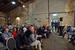 Eine Gruppe Menschen sitzt auf Stuhlreihen in einem alten Saal mit halbverputzten Wänden und Holzdecke. diskutiert. Ein Mann in der ersten Reihe spricht in ein Mikro. 